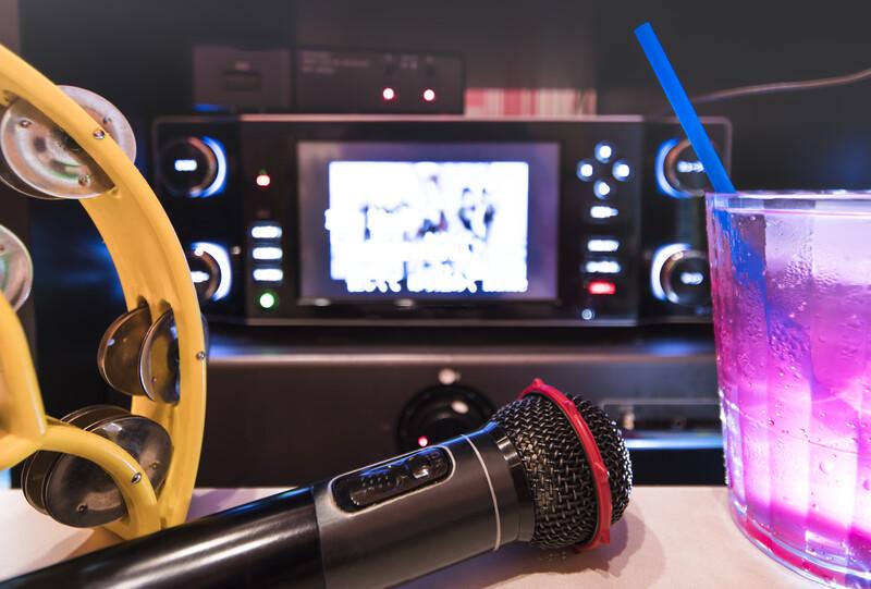 Black microphone in karaoke club, with remote controller, melon and strawberry soda drinks, yellow tambourine and screen for singing music on stage pa